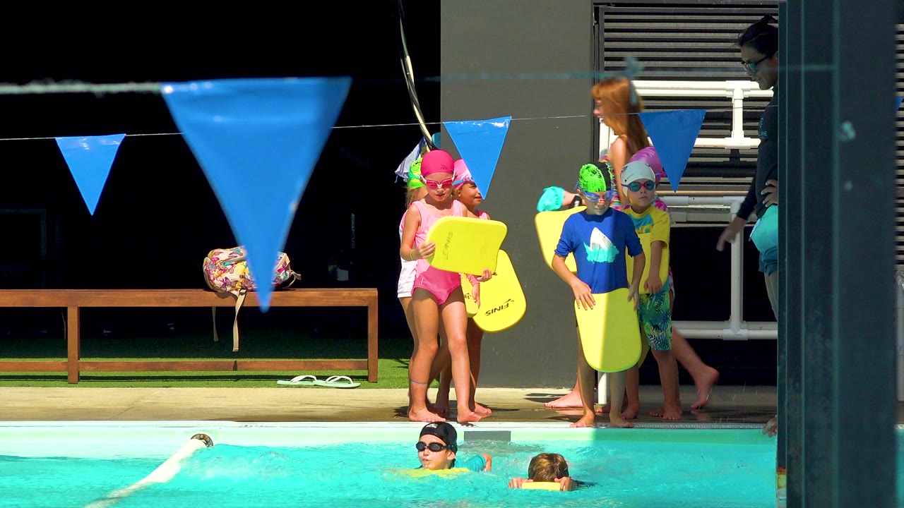 Children in colorful swim caps and goggles stand at a Cabo San Lucas pool edge with yellow kickboards, blue flags in the foreground, and swimmers partly visible in the water.