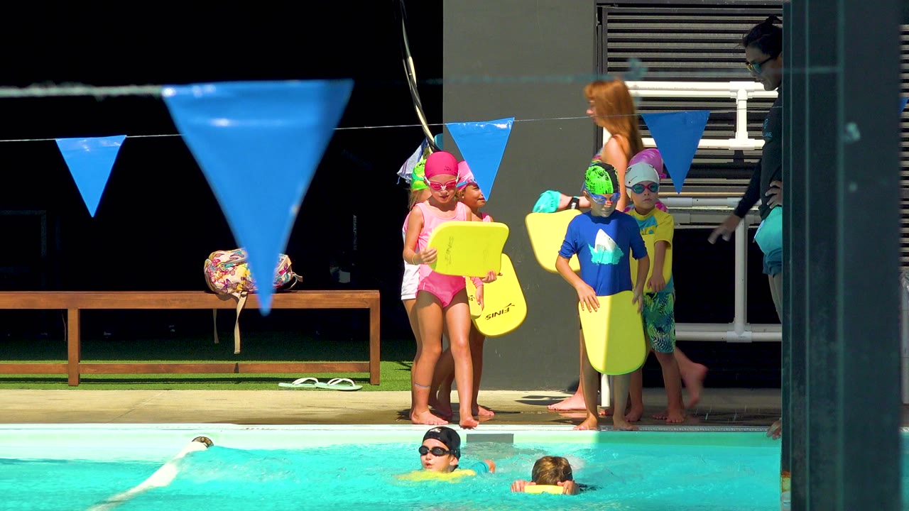 Children with kickboards wait near the pool while an adult stands behind them.