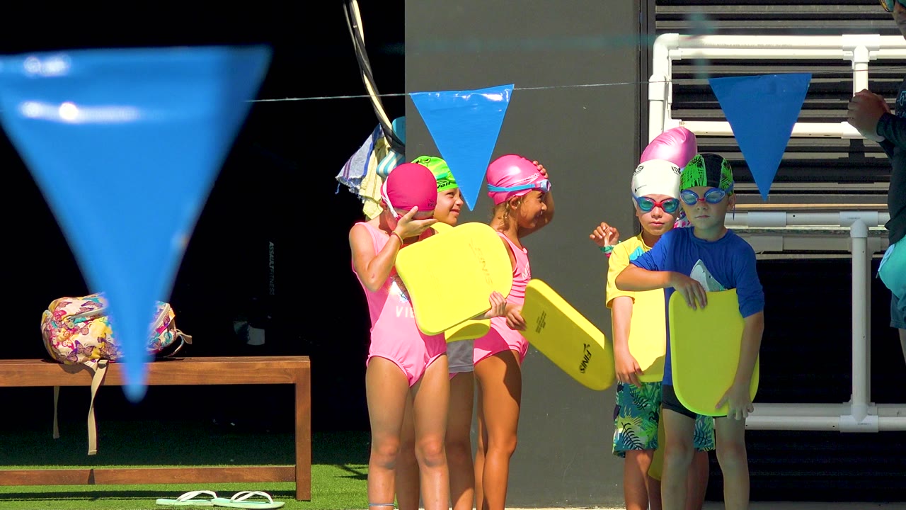 Children in Cabo San Lucas, Mexico stand near a pool with yellow kickboards, bright swim caps, goggles, and a large blue pennant flag crossing the foreground.