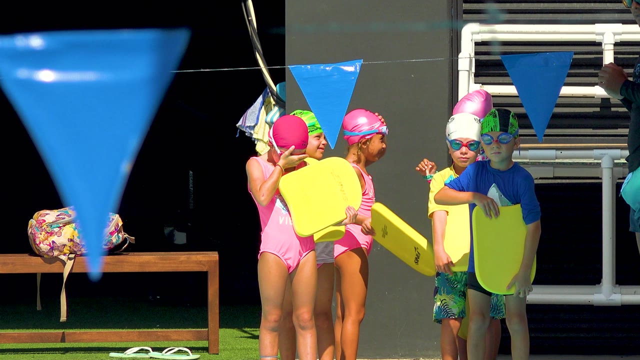 Children in swimwear cluster with kickboards beside pool flags.