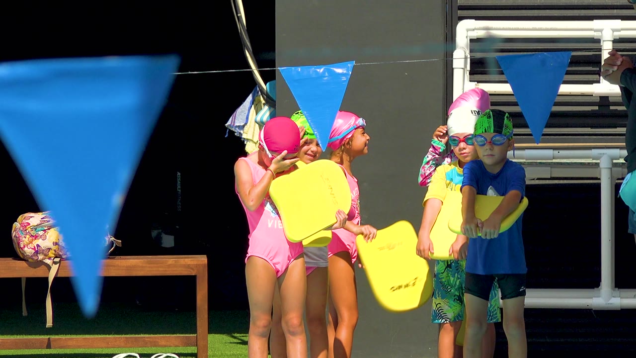 Several children in Cabo San Lucas, Mexico adjust yellow kickboards while one child in a blue shirt stands front-facing beneath the blue pool flags.