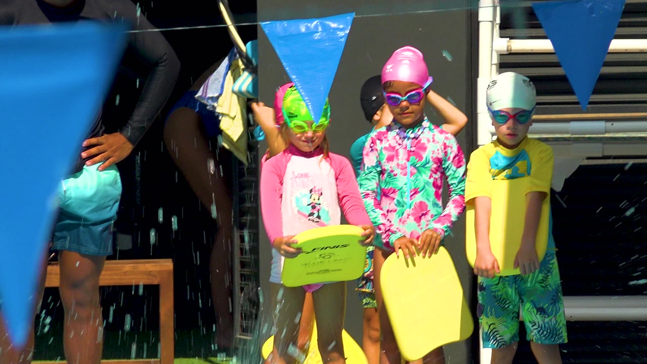 Children in colorful swim caps and goggles stand at a Cabo San Lucas pool edge with yellow kickboards, blue pennant flags, and strong sunlight across their swimwear.