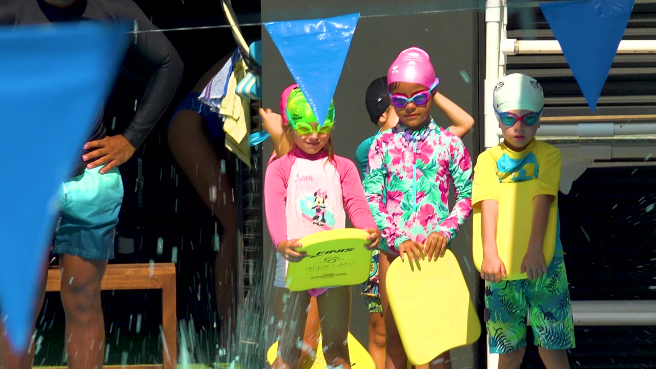 Children stand in a row with kickboards as water splashes in front of them.