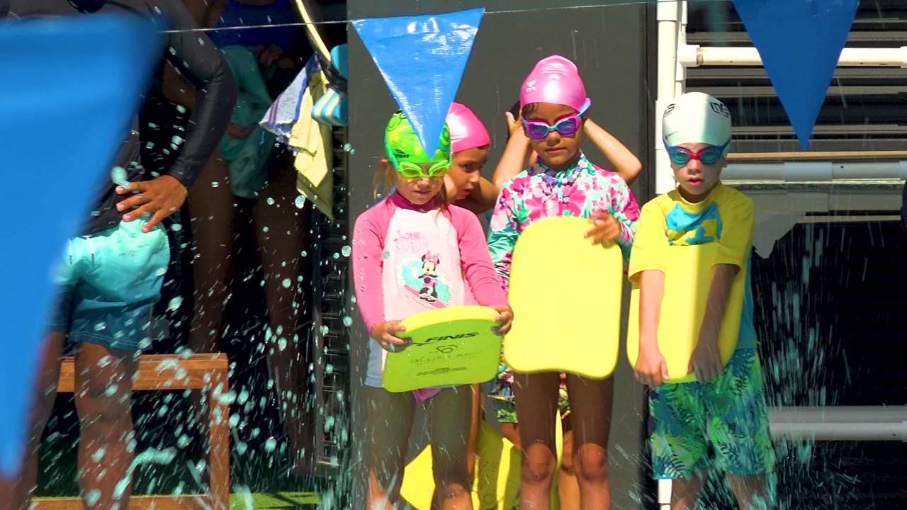 At 0:02, pool spray falls across the foreground as children in swim caps and goggles hold yellow kickboards near the pool edge in Cabo San Lucas.