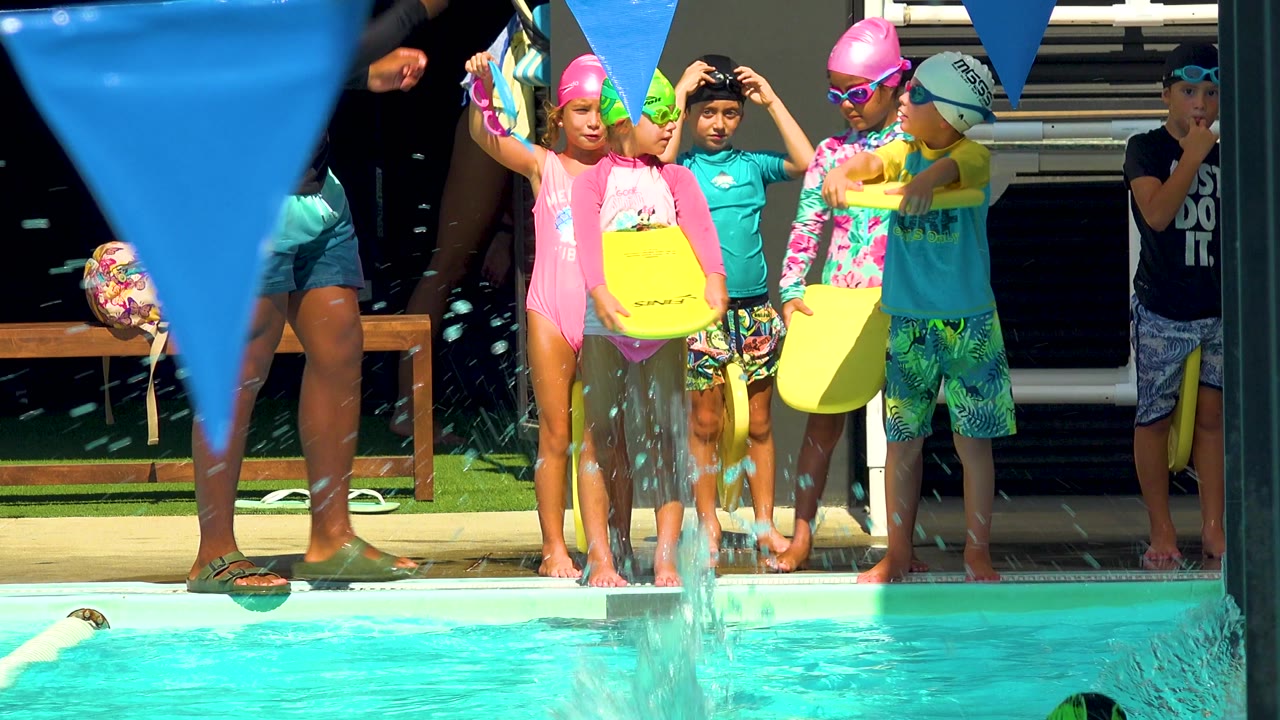 Children in swim caps and bright rash guards stand at a pool edge in Cabo San Lucas, Mexico, holding yellow swim planks as water splashes in front of them.