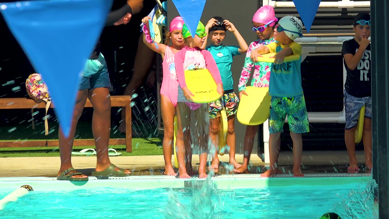 Children wait with swim planks near splashing pool water.