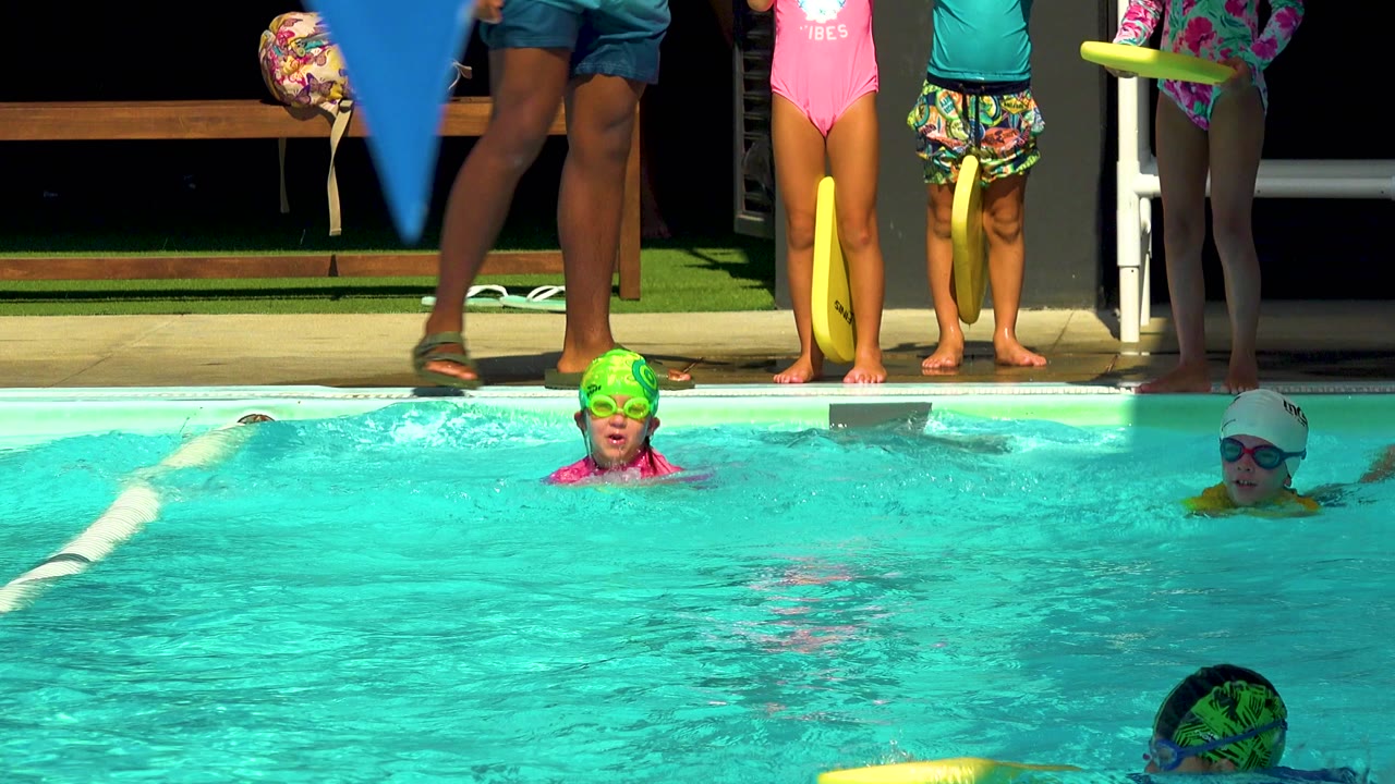 At 0:00 in Cabo San Lucas, children in swim caps and goggles are spread across a turquoise pool while cropped adults and waiting children stand on the deck.