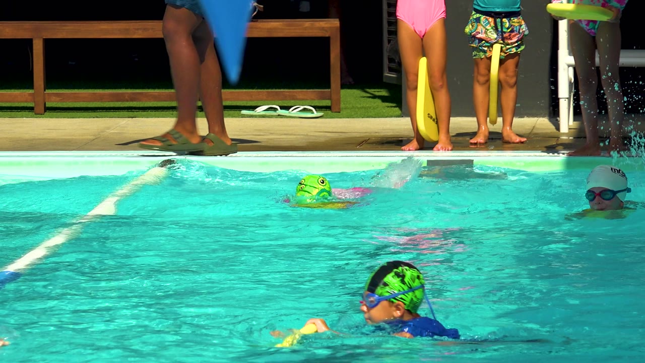 At 0:02 in Cabo San Lucas, multiple children move through the pool, including a swimmer in a green cap near the center and a swimmer in a white cap at the right side.