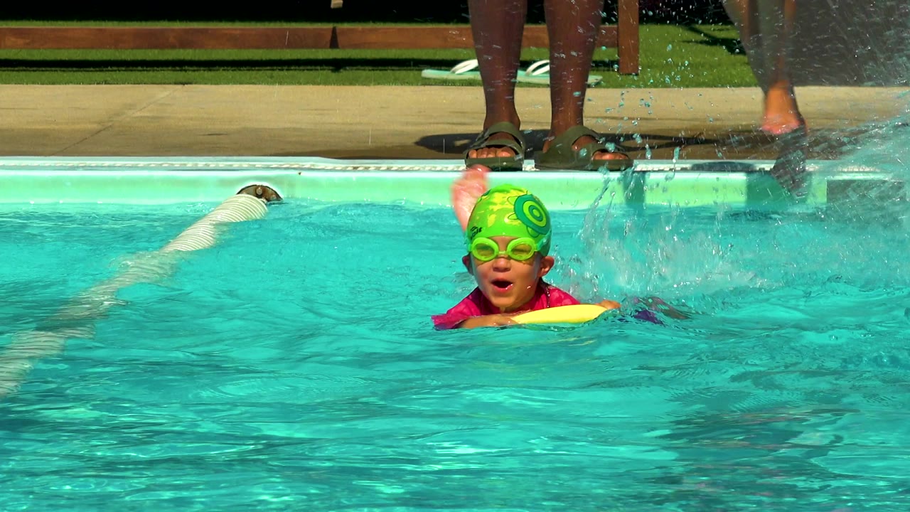 In Cabo San Lucas, Mexico, a child in a green swim cap and goggles holds a yellow foam float while moving through bright turquoise pool water.