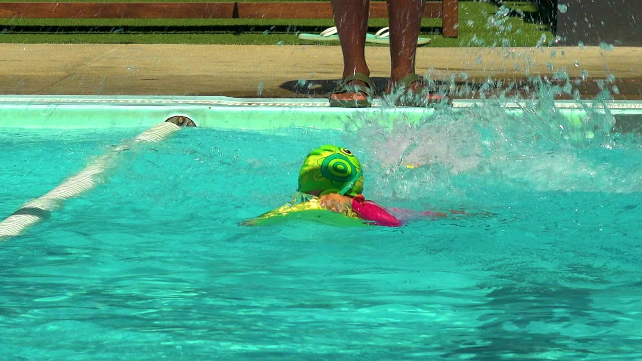 A child in a green swim cap leans forward on the yellow float while kicking, with a large splash rising near the pool edge in Cabo San Lucas.