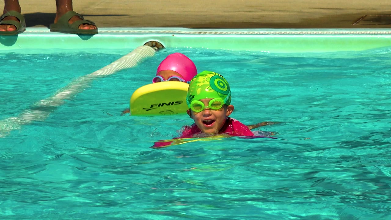 In Cabo San Lucas, Mexico, two children wearing bright swim caps and goggles hold yellow kickboards near a lane rope in turquoise pool water.