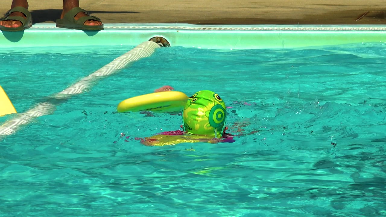 In Cabo San Lucas, Mexico, a child in a green swim cap moves across the pool with a yellow kickboard floating nearby and the lane rope angling behind.