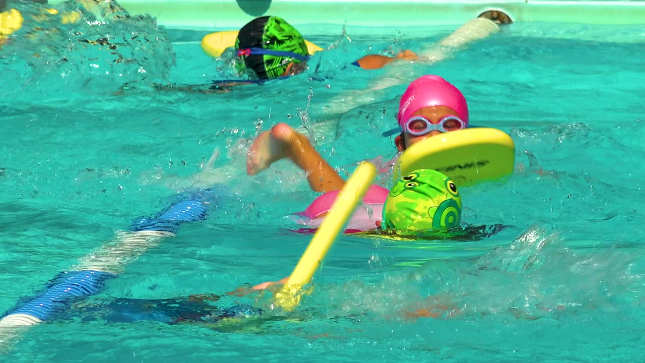 Two children swim close together with kickboards in turquoise pool water.