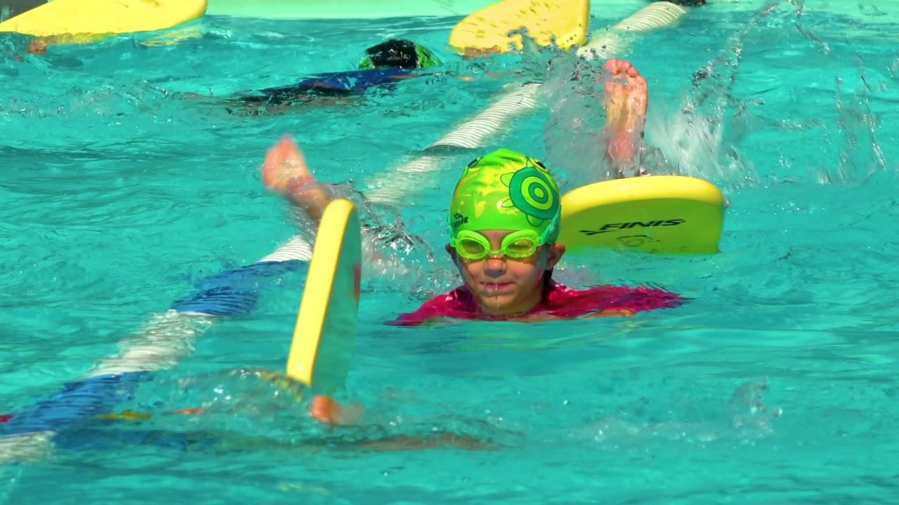 A child in a green swim cap and goggles faces the camera in a Cabo San Lucas pool as another child kicks beside a yellow kickboard near the lane rope.