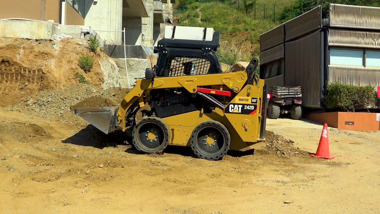 Yellow skid-steer loader with a front bucket on tan dirt at a Cabo San Lucas construction site, with nearby buildings, a hillside, an orange cone, visible CAT branding, and no visible people.