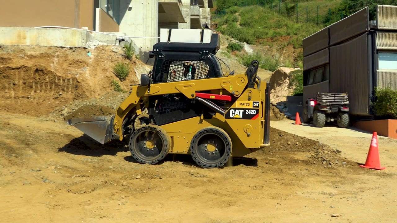 Yellow CAT-branded skid-steer loader at the Cabo San Lucas construction area, framed against exposed soil, a driveway or ramp, a dark trailer or structure, and no visible people.