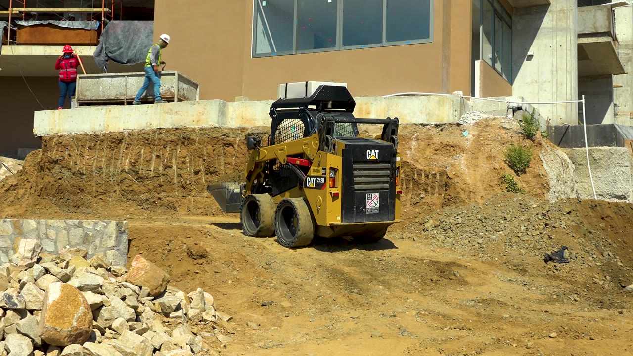 The loader remains centered on the dirt work area in Cabo San Lucas, Mexico, while workers stand along the upper concrete edge beside a tan building facade.