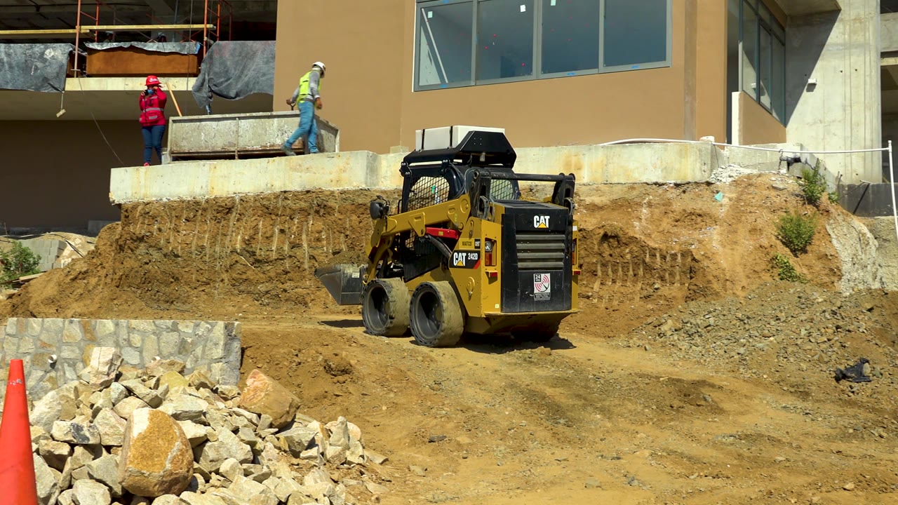 A yellow loader works below an exposed dirt bank in Cabo San Lucas, Mexico, with an orange traffic cone and pale rocks visible in the foreground.