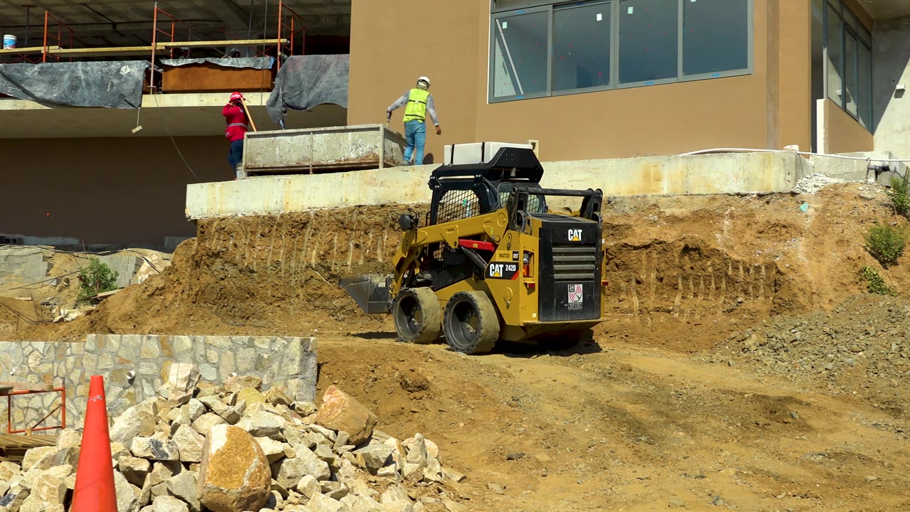 Skid steer loader below a building under construction with workers on the ledge.