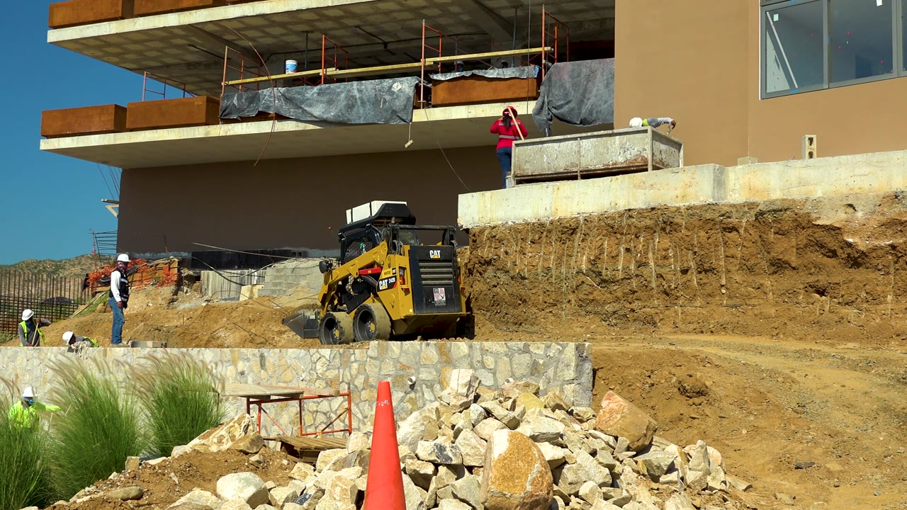 Workers in hard hats are visible around a yellow skid steer near a rock wall and exposed soil at the Cabo San Lucas build site.