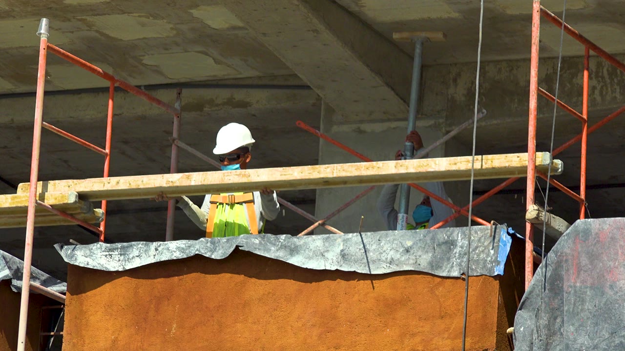 A construction worker in Cabo San Lucas stands behind an orange platform and red scaffold rails while a long wood plank crosses the frame near the start of the clip.