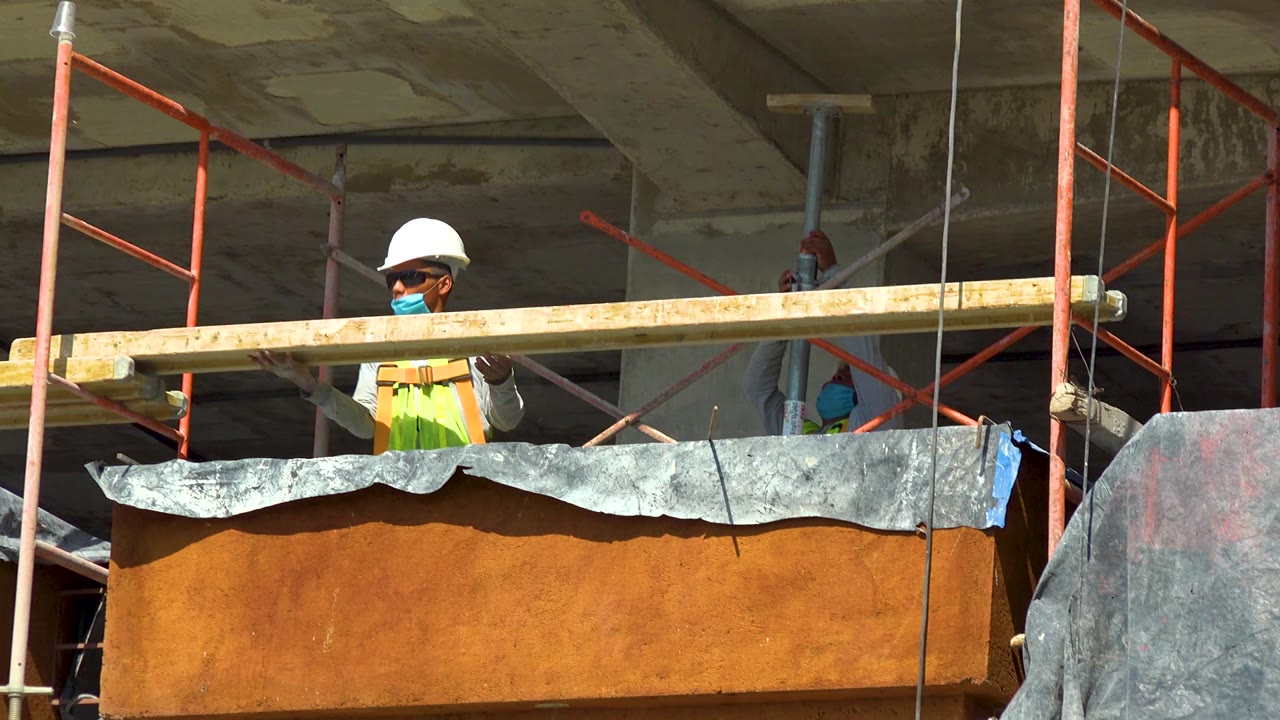 A hard-hatted worker in Cabo San Lucas handles the long wood piece behind an orange platform, with red scaffold pipes and a gray hanging tarp around the work area.