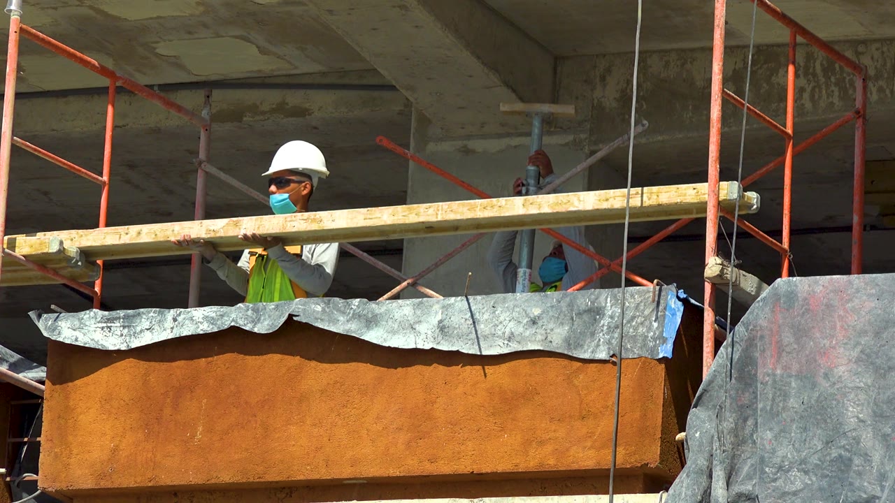 Worker turns beside a long plank on red scaffolding.