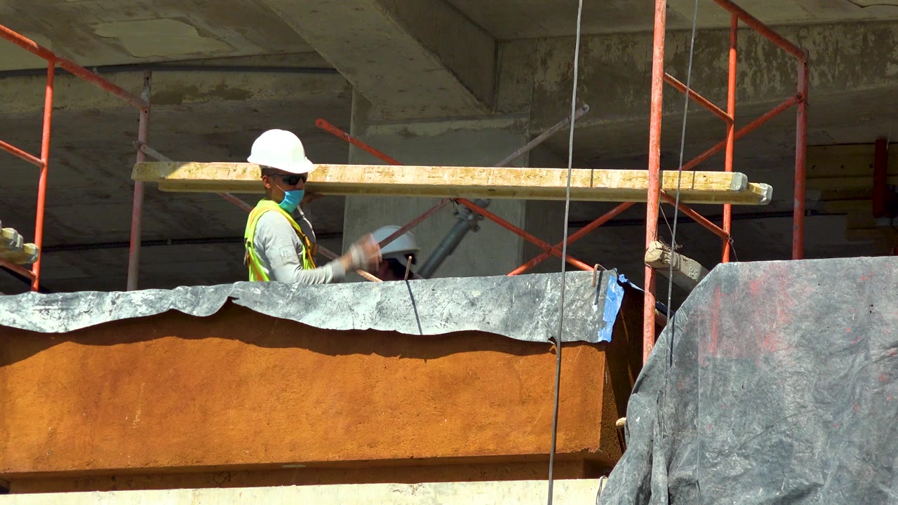 In Cabo San Lucas, Mexico, a construction worker in a white hard hat, blue mask, and neon vest stands beside a long wood beam on red scaffolding above orange-brown formwork.