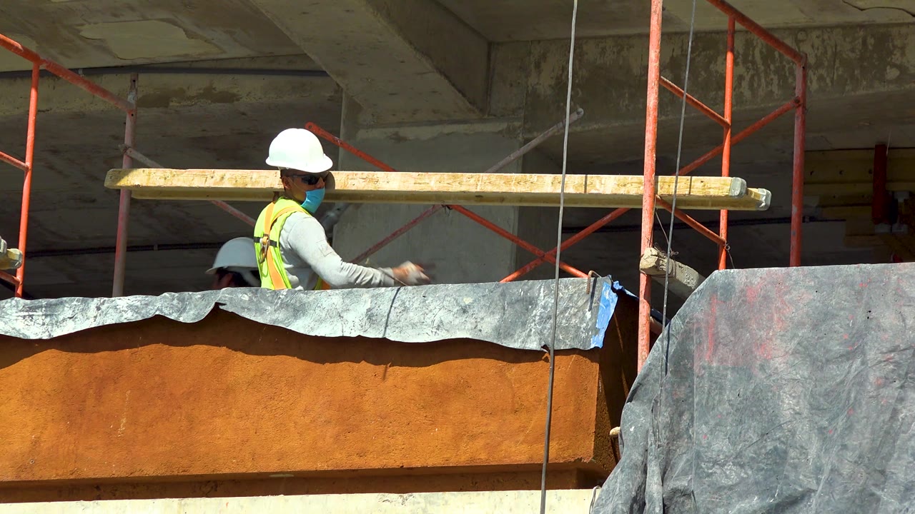 In Cabo San Lucas, Mexico, the worker turns near the long beam, with red scaffolding behind him and a second white hard hat partly visible behind the foreground barrier.