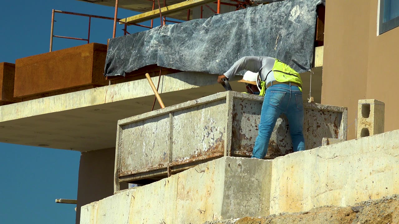 In Cabo San Lucas, the worker remains bent over the ledge with his back toward the camera, framed by scaffolding, black protective sheeting, and a tan wall.