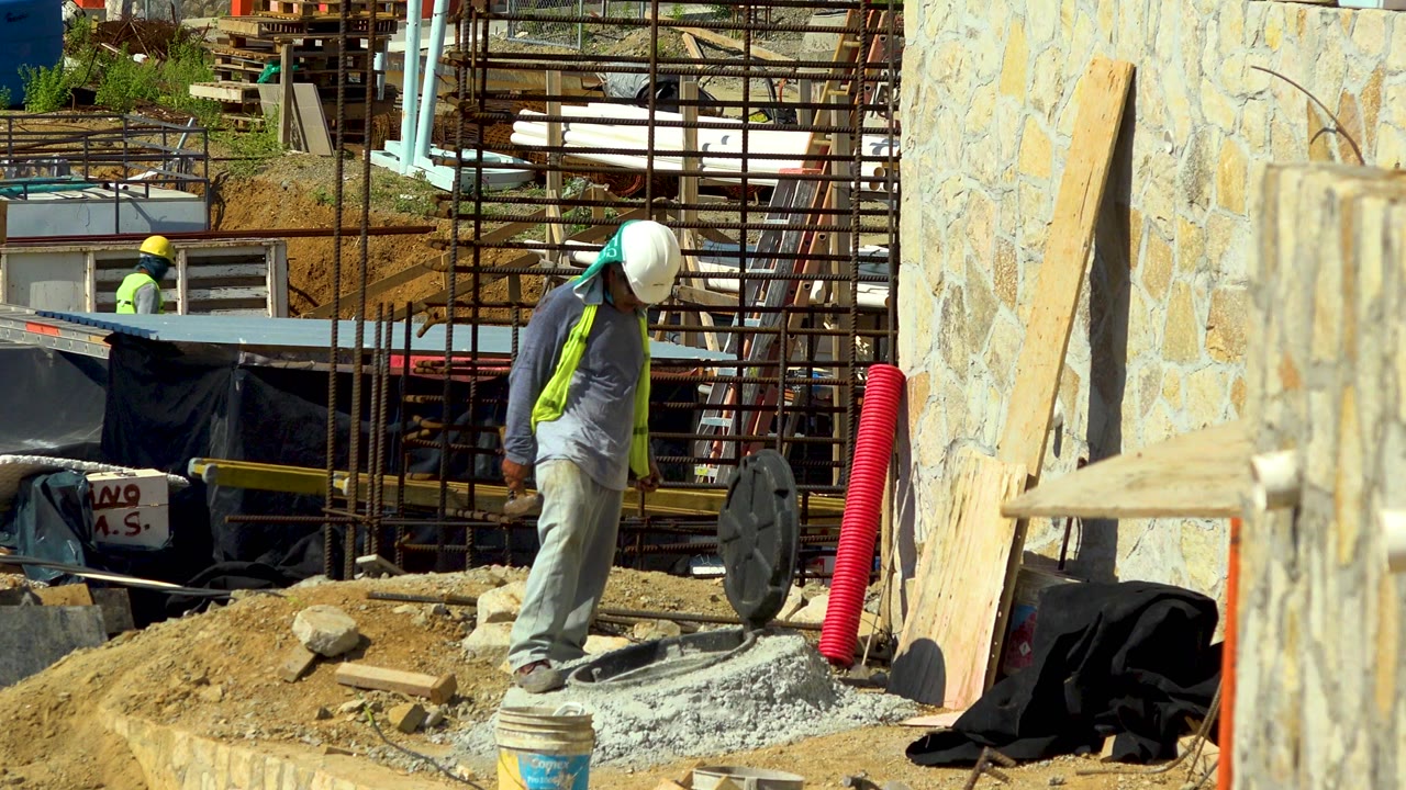 A construction worker in a white hard hat or cap stands on gravel at a Cabo San Lucas worksite, with rebar, scaffolding, a stone wall, and a red corrugated pipe around him.