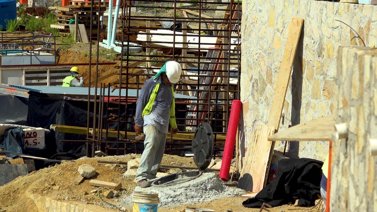 The worker remains near the gravel mound at the Cabo San Lucas construction site, with the red corrugated pipe, stone wall, wood planks, and scaffold lines framing the scene.