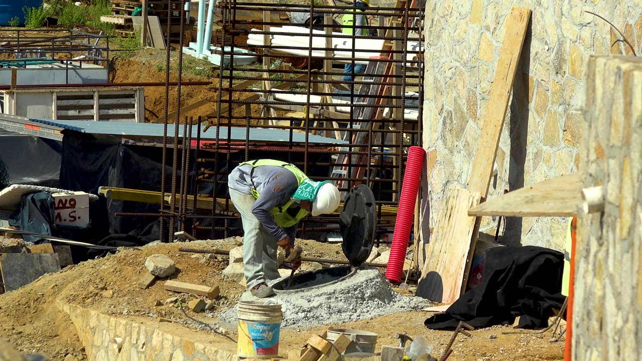 A helmeted construction worker bends over a patch of wet concrete at an outdoor construction site in Cabo San Lucas, Mexico, with sand, buckets, rebar, scaffolding, and a stone wall around him.