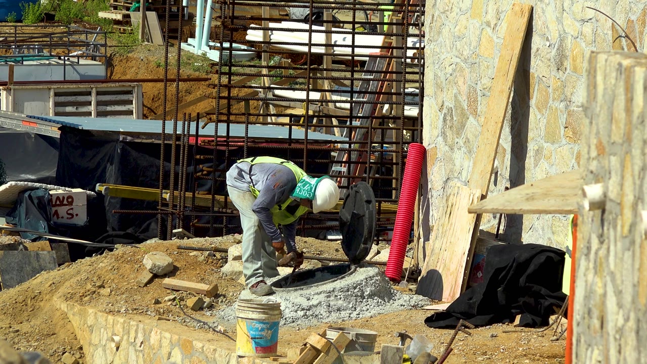 A construction worker in a hard hat leans lower over the concrete in Cabo San Lucas, Mexico, with the red corrugated pipe and pale stone wall more prominent on the right side of the frame.