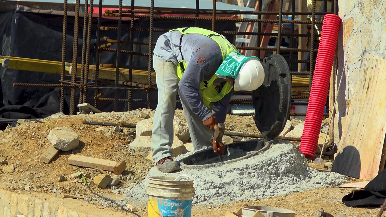The worker stays bent over the concrete manhole in Cabo San Lucas, with a tall red corrugated conduit and dark round cover visible to his right.