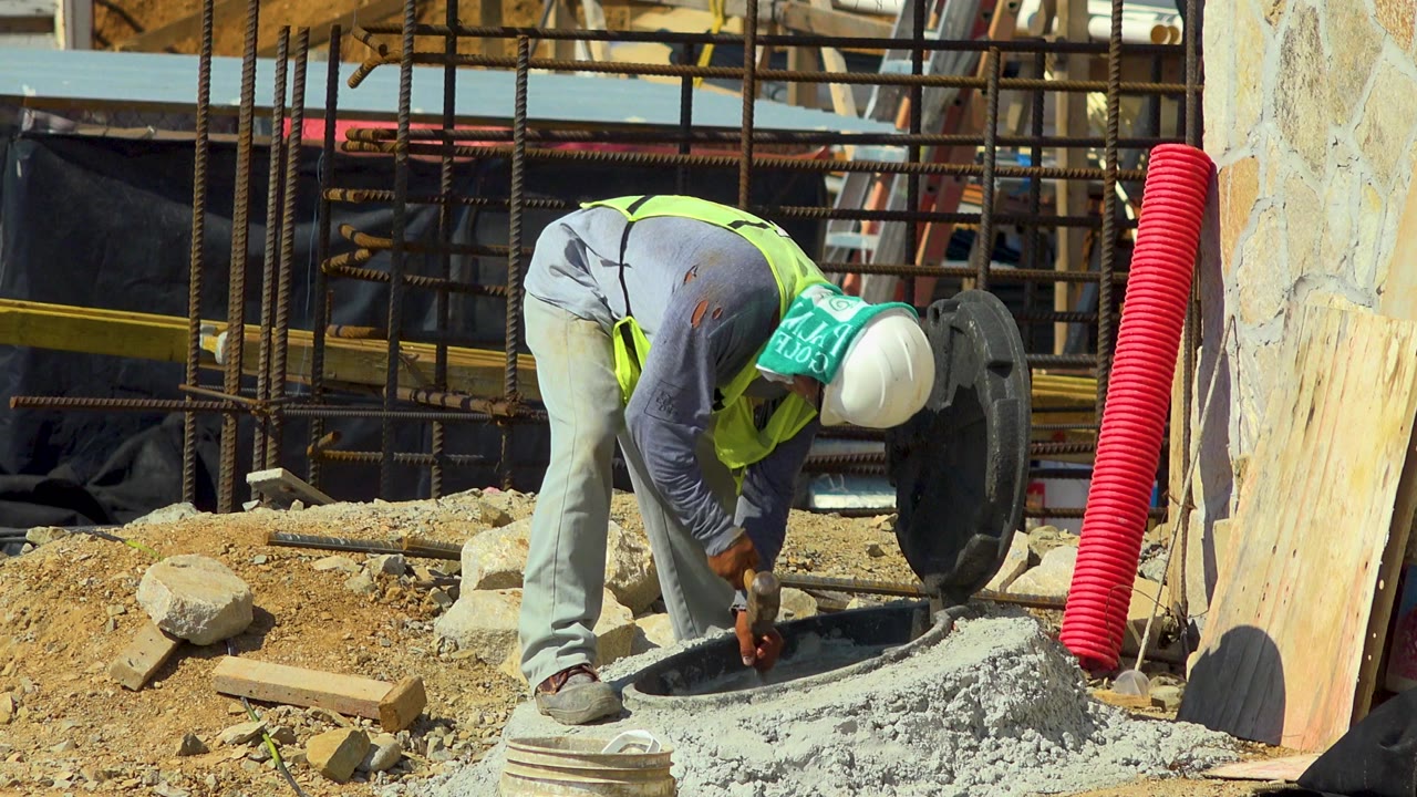In Cabo San Lucas, Mexico, a hard-hat construction worker bends over a round concrete opening while chiseling, with dirt, rebar, lumber, and a red corrugated pipe behind him.