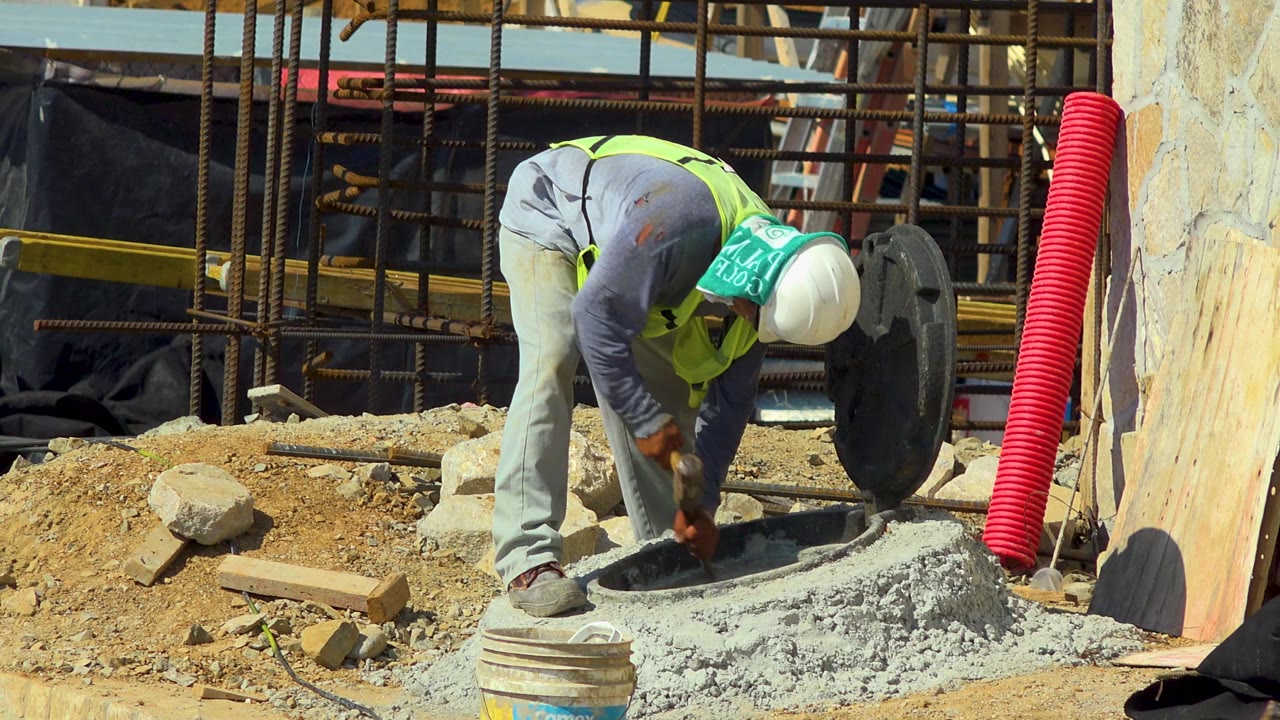 In Cabo San Lucas, Mexico, the worker leans deeper over the concrete hole, with the red corrugated pipe more prominent on the right and rubble scattered across the dirt foreground.