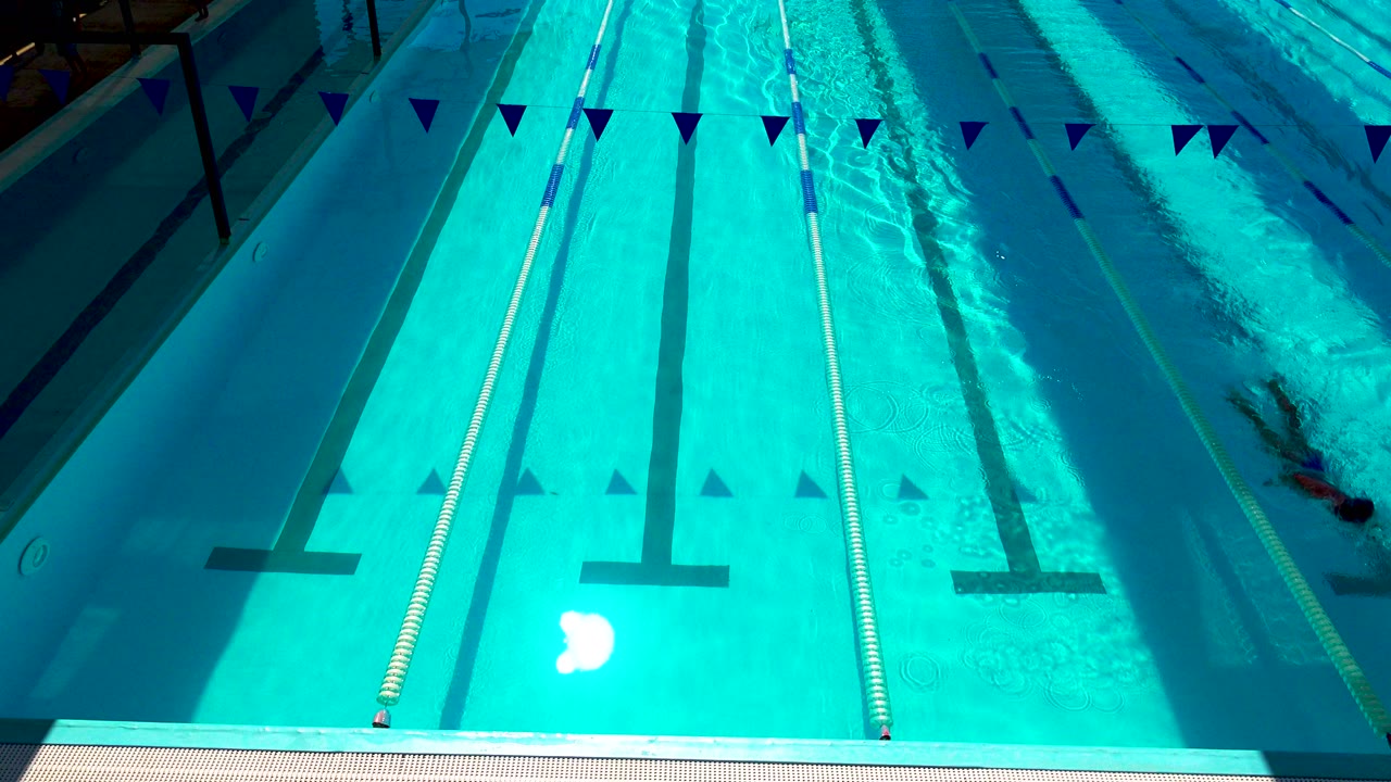 Wide overhead view of a sunlit blue lap pool in Cabo San Lucas with lane ropes, dark lane markings, flags across the top of frame, and no visible people.