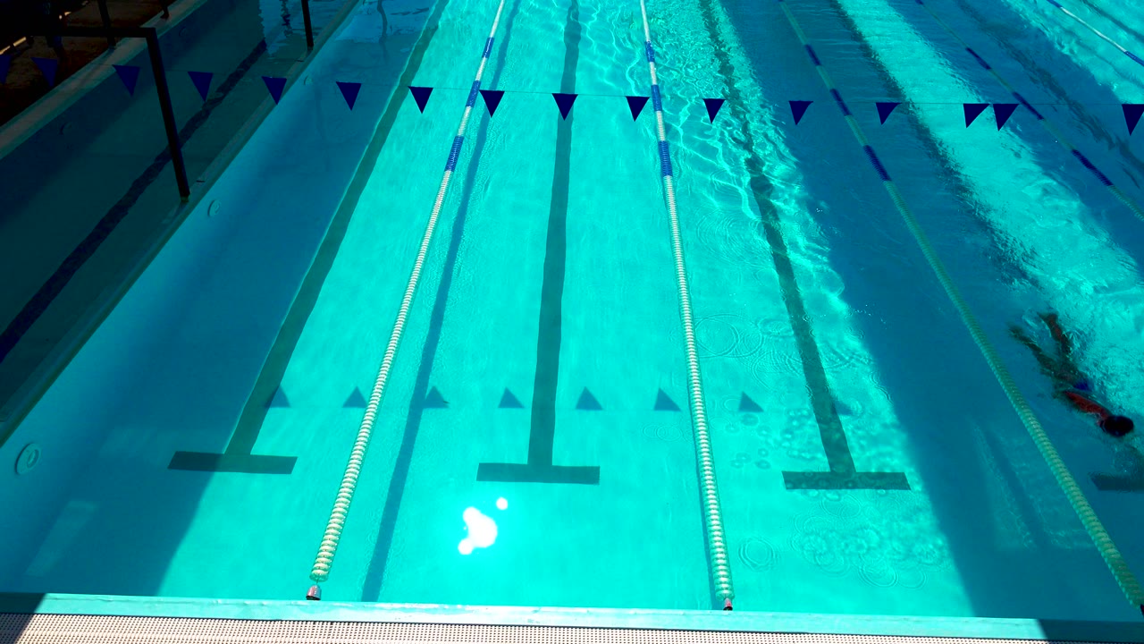 High-angle view of the Cabo San Lucas pool with bright turquoise water, lane ropes, overhead flags, long shadows, and a swimmer just visible near the right edge.