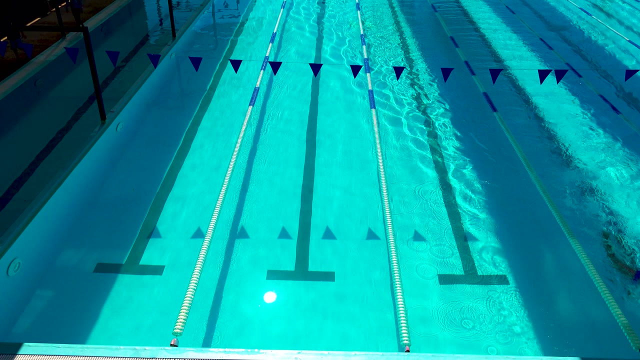 The pool remains framed from above in Cabo San Lucas, showing lane ropes, dark markings, flags, sun glare near the lower center, and no visible people in this moment.