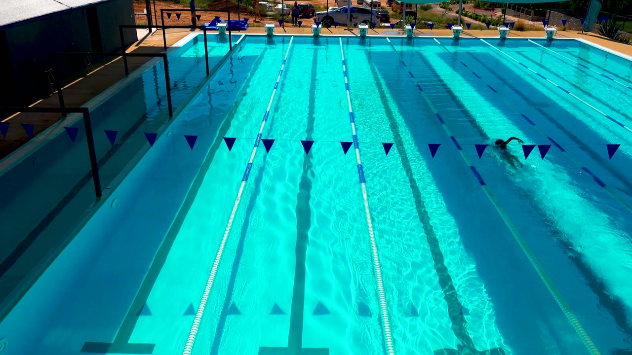 A distant swimmer is visible near the right lanes of the Cabo San Lucas outdoor pool, with blue pennant flags across the foreground and strong sunlight on the water.
