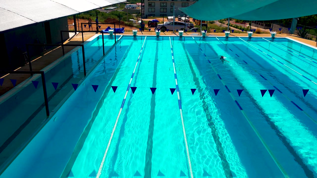 At 0:00, a Cabo San Lucas resort lap pool is seen from above with turquoise water, blue pennants, lane ropes, shade sails, and one swimmer near the far lanes.