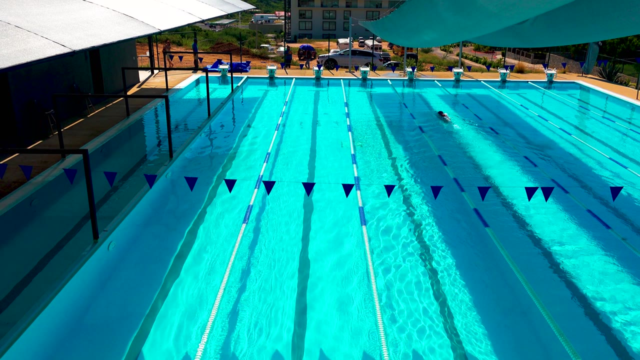 At 0:02, the elevated view holds on the Cabo San Lucas pool as the lone swimmer appears farther across the upper lanes, with a building and parked cars beyond the deck.