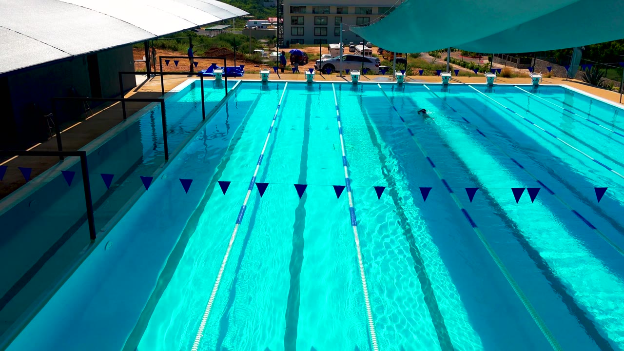 Turquoise resort pool with lane lines, overhead pennants, shade sails, and a swimmer near the far edge.