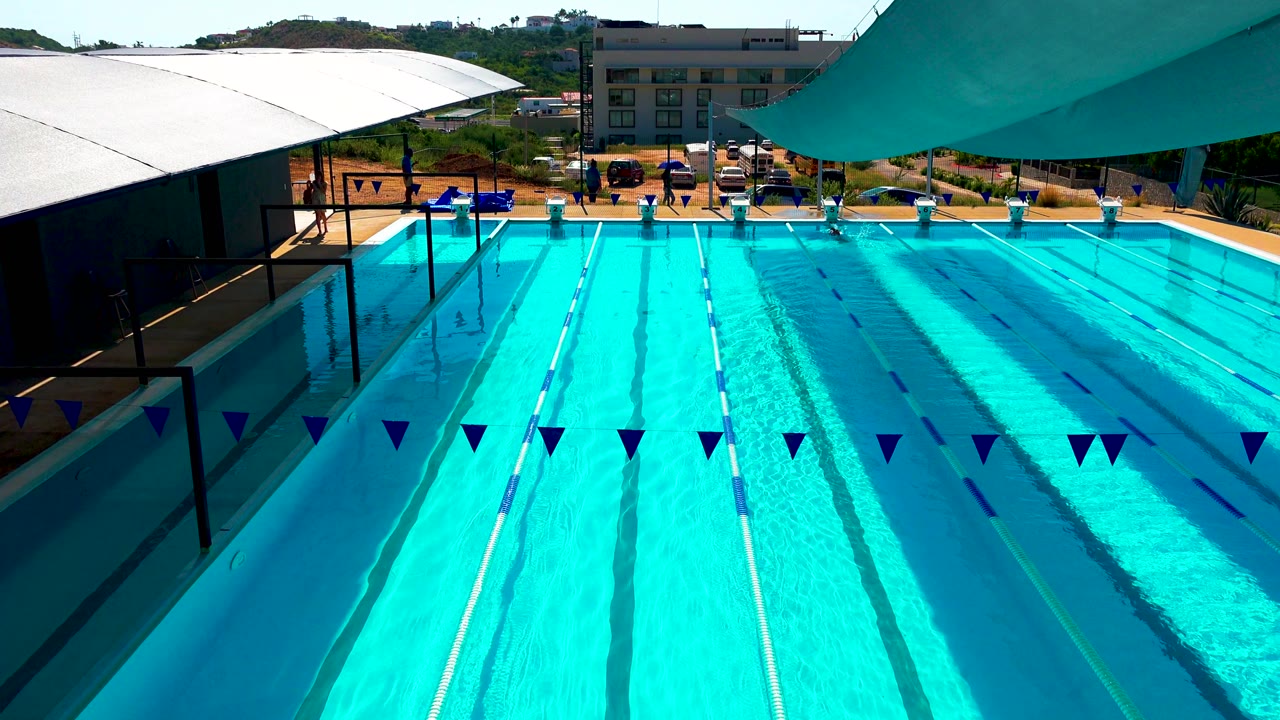 Cabo San Lucas outdoor resort pool at the start of the clip, with bright blue lap lanes, triangular pennant flags across the foreground, white and teal shade canopies, and small people on the far deck.