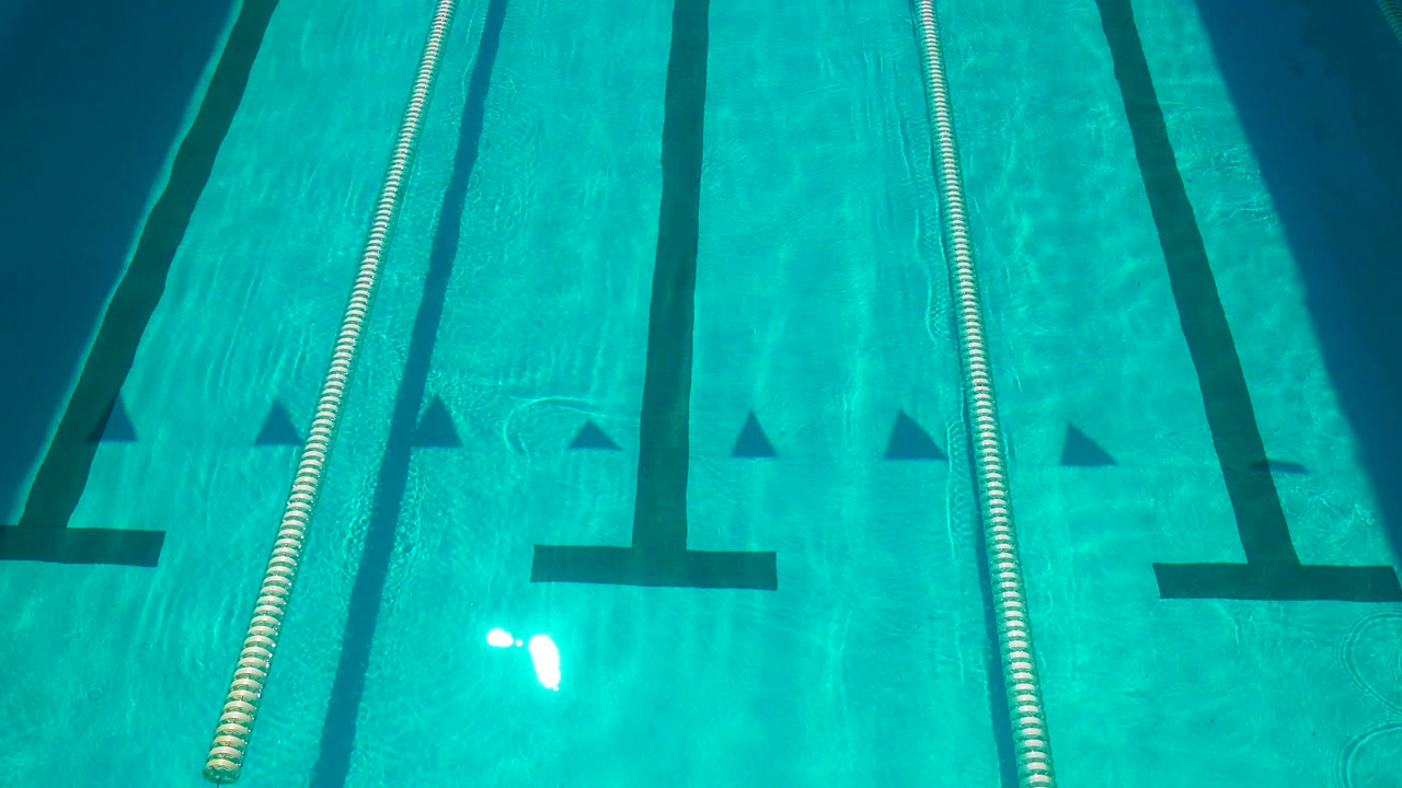 Sunlit Cabo San Lucas pool lane with a white floating rope, dark T-shaped floor markings, small triangular markers, and no visible people.