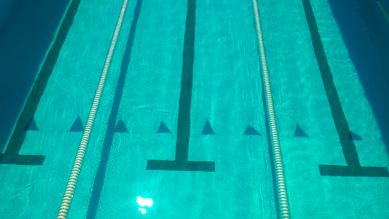 Cabo San Lucas competition pool surface with a lane rope along the left edge, dark black lane markings across blue water, bright light reflection, and no visible people.