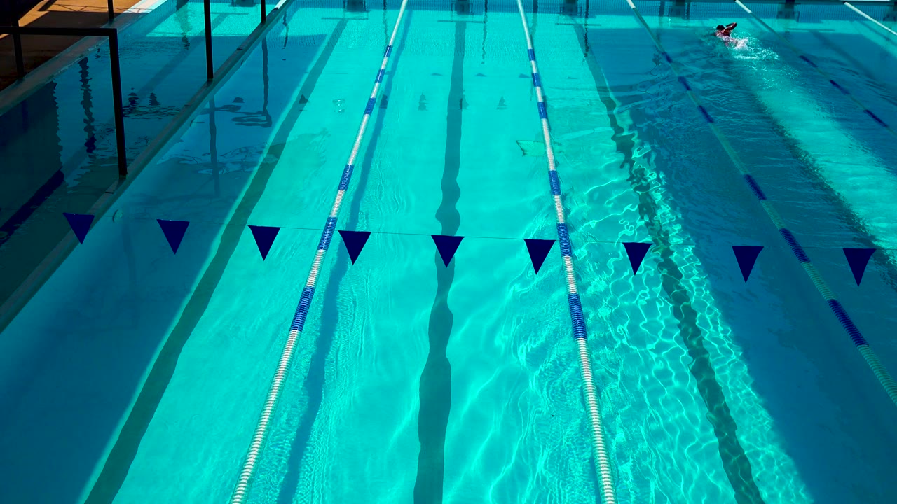 Cabo San Lucas outdoor lap pool seen from above, with turquoise water, lane dividers, dark blue pennants in the foreground, and a small swimmer near the far right.
