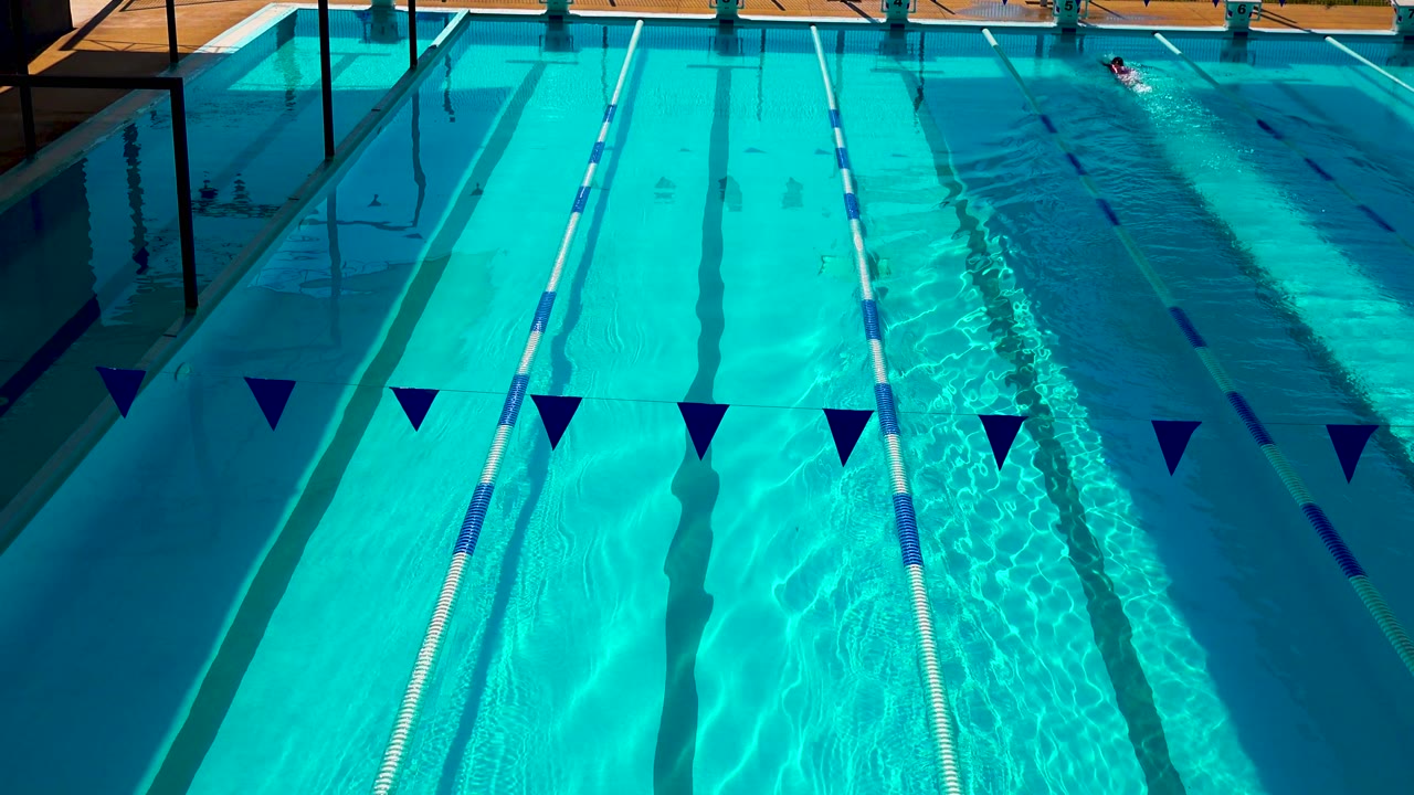 Cabo San Lucas outdoor pool with lane dividers, bright blue water, diagonal shadow bands, foreground pennants, and a small swimmer near the far end of the right lanes.