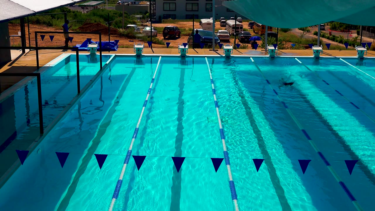 Wide Cabo San Lucas swimming pool frame with dark lane lines, blue-and-white ropes, triangular flags, shade across the top edge, and a swimmer small in the upper right of the water.
